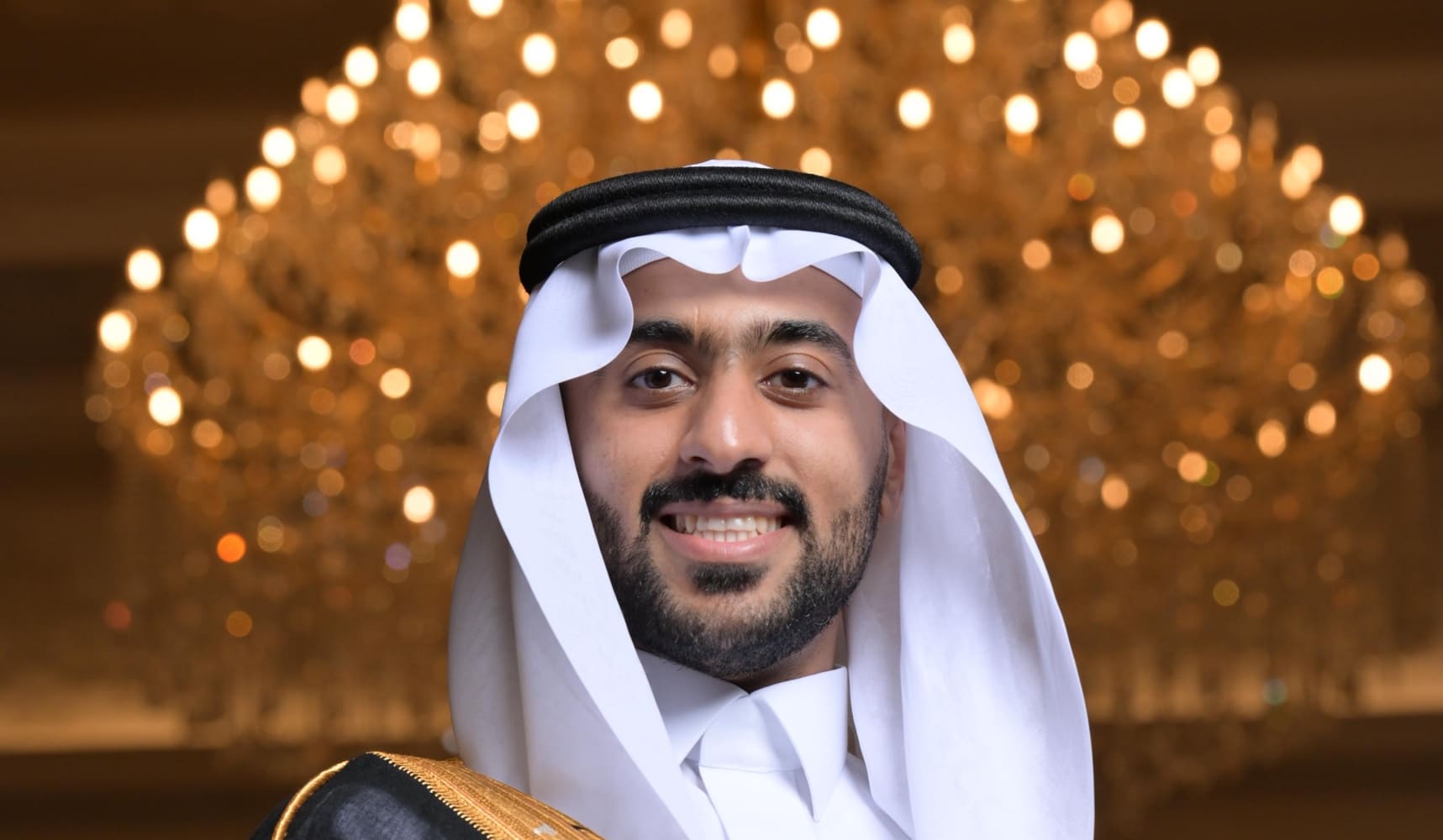 Abdulaziz Bin Odah in a white thobe, shemagh and black bisht with gold trim, smiling, in front of a lit chandelier